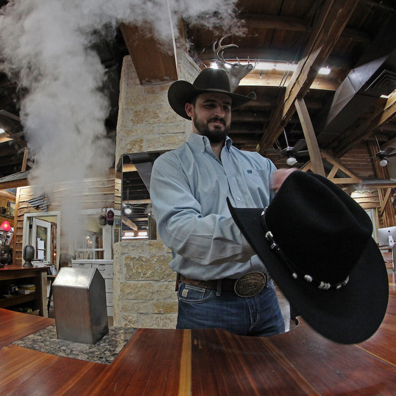 Hatter in a black hat steaming a felt hat at the shop counter